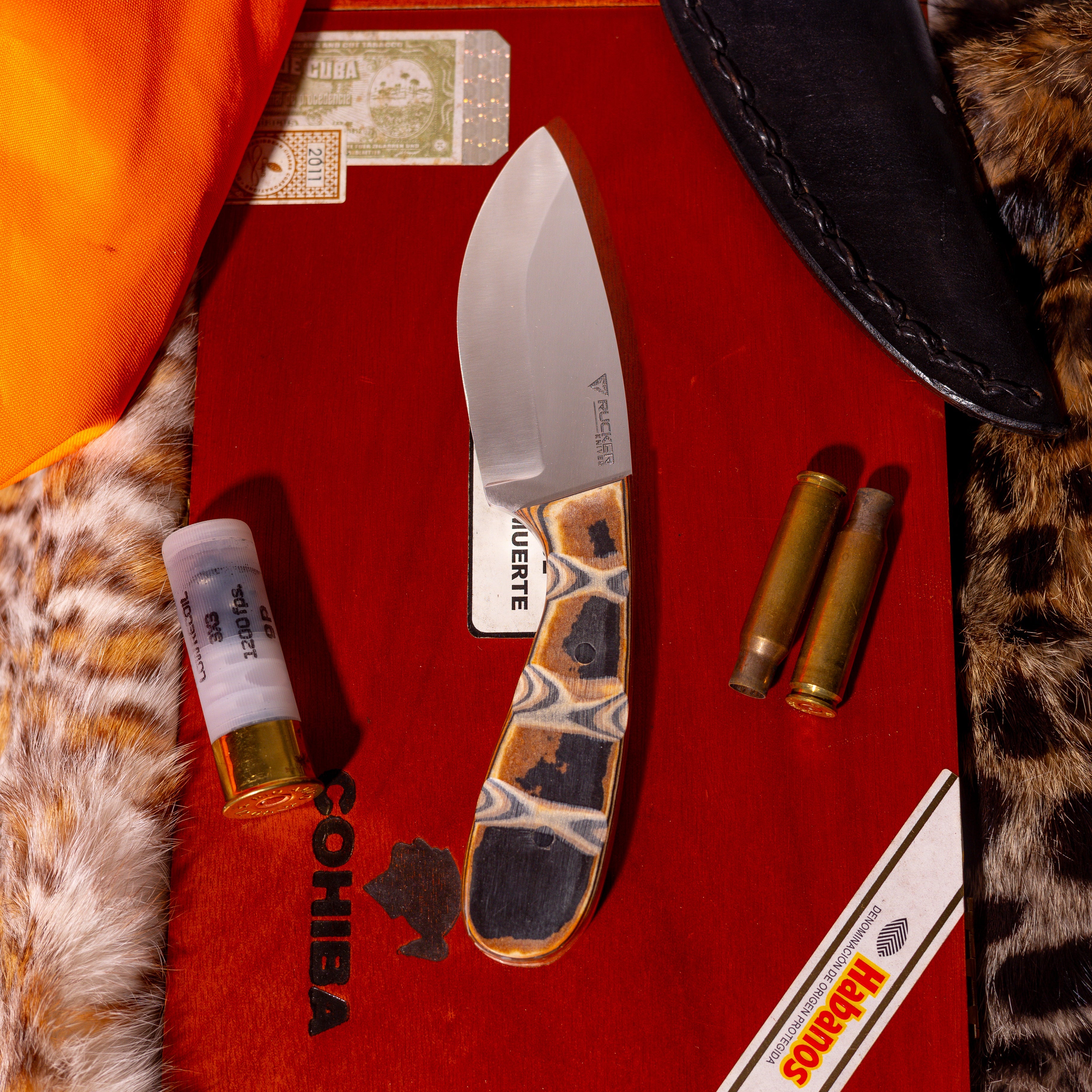 Rucker Knives Griz-Skinner Knife staged background on cigar box, leopard print, and leather sheath